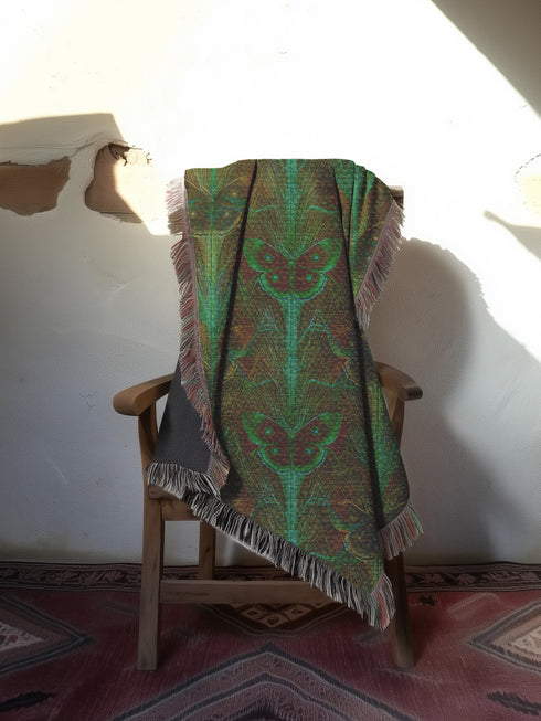 A woven cotton throw featuring an intricate luna moth design in deep green and russet tones, framed by soft fringe.